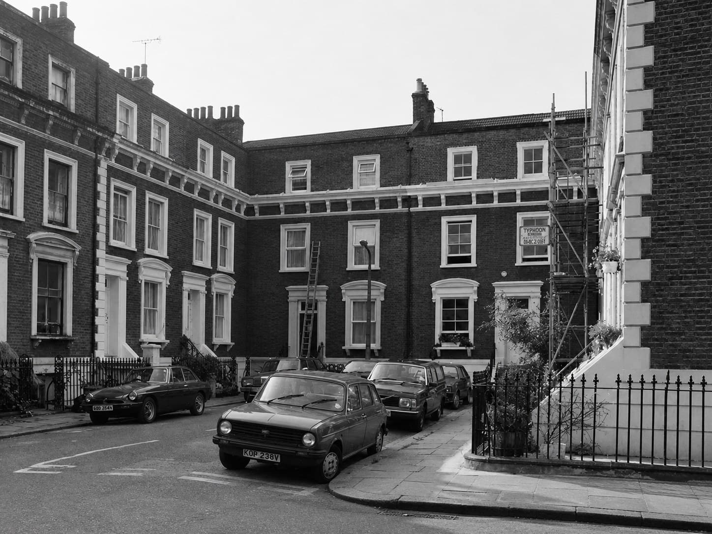 Black-and-white photograph of a Victorian terrace in Hackney showing scaffolding and restoration work in progress, with multiple period vehicles parked along the street, photographed in the early 1980s.